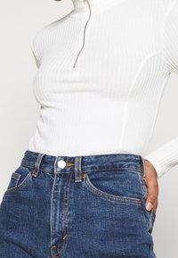 Wit geribd shirt met lange mouwen en een halve rits, gecombineerd met hoge taille blauwe spijkerbroek met knoopsluiting en contrasterende stiksels.