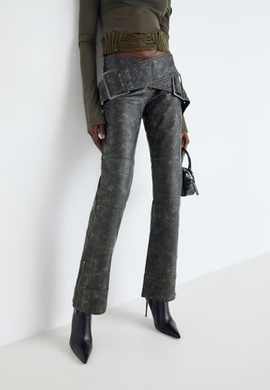 Person iført slidte mørkegrå læderbukser med brede bælte detaljer, olivengrøn cropped top, sorte spidsede høje hæle støvler og holder en lille taske.