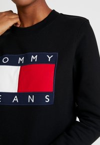 Musta collegepaita, jossa on värikäs logo punaista, valkoista ja tummansinistä suorakulmamaista elementtiä sekä valkoista brodeerattua tekstiä "TOMMY JEANS."