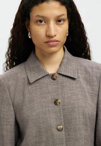 Camicia grigia in lino con colletto a punta e quattro bottoni circolari marroni lungo la parte anteriore, taglio strutturato e tessuto testurizzato.