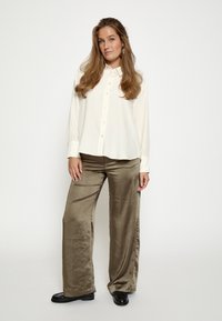 Kreemipehme pluus nööpidega, mugav lõige ja pikad varrukad, koos oliivroheliste satiinist laiade pükstega, lõpetatud mustade loaferitega.