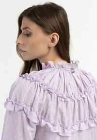 Lichtpaars blouse van textuurstof, met geruffelde accenten en een geraffineerde halslijn. Gedoteerd patroon door de gehele blouse.