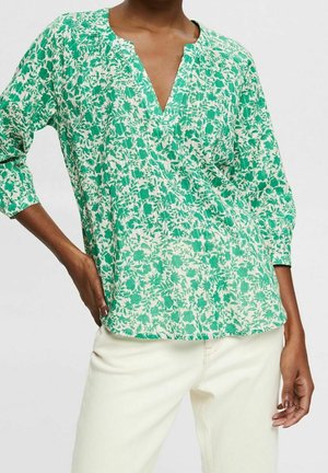 Kvinde iført en grøn og hvid blomsterbluse med V-hals og trekvartærmer, kombineret med off-white bukser mod en ensfarvet baggrund.