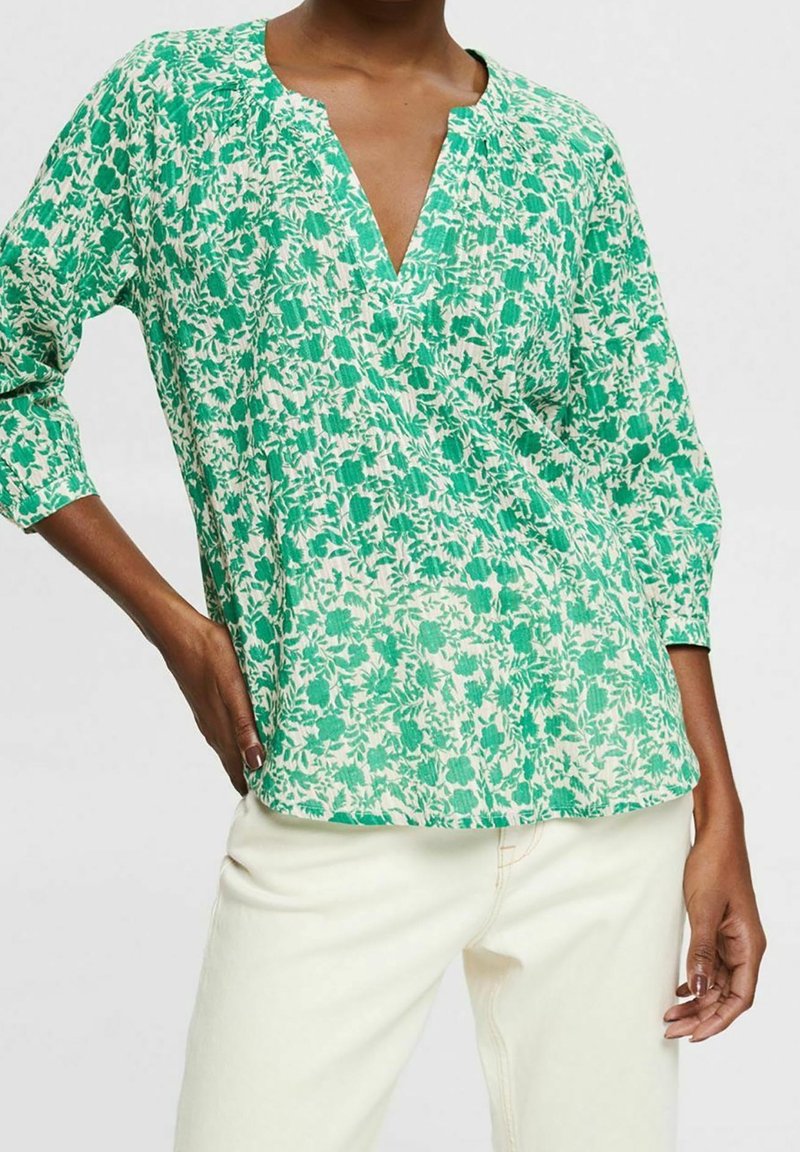 Femme portant un chemisier à fleurs vert et blanc avec un col en V et des manches trois-quarts, associé à un pantalon blanc cassé sur un fond uni.