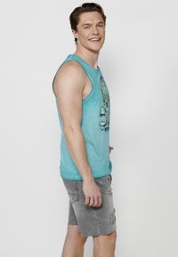 Turkos linne med grafiskt tryck och grå slitna shorts. Tyget är lätt; passformen är avslappnad och ledig.