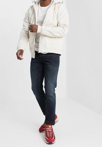 Man wearing white hoodie, white t-shirt, dark jeans, and red sneakers walking against plain white background.