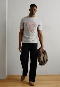 Grey short-sleeve t-shirt with red circular logo, dark wide trousers, and beige shoes. Holding a light brown jacket. Simple, casual look.