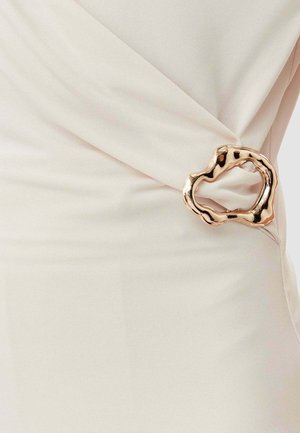 Abito color crema con design drappeggiato, tessuto texturizzato e una fibbia metallica dalla forma organica in vita.