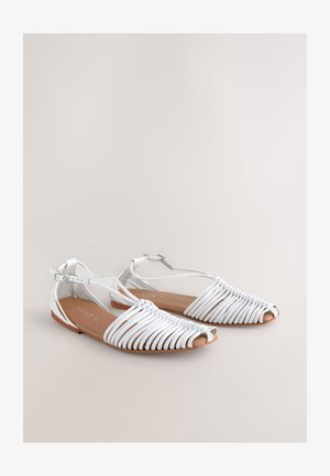 Białe sandały na paski z odkrytymi palcami, o plecionej konstrukcji, z paskiem wokół kostki z klamrą i płaską brązową podeszwą. Gładka faktura, styl casualowy.