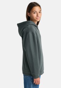 Teenager mit einem dunkelgrauen Reißverschluss-Hoodie, dessen Kapuze heruntergeklappt ist, kombiniert mit blauen Jeans, steht seitlich und blickt in die Kamera.