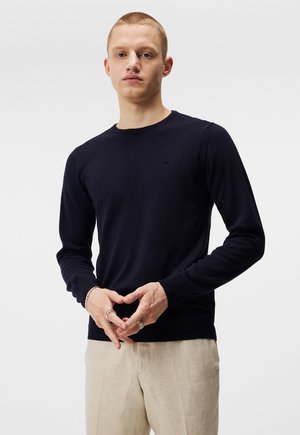 Navyblaues Strickpullover mit Rundhalsausschnitt und langen Ärmeln, ausgestattet mit einem kleinen schwarzen Logo auf der Brust sowie gerippten Bündchen und Saum.