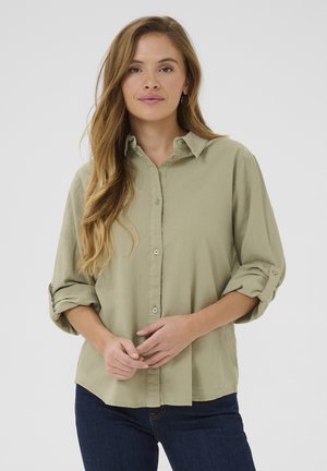 Hellgrüne, knöpfbare Bluse mit weicher Textur, ausgestattet mit einem Kragen und hochgerollten Ärmeln, kombiniert mit dunkelblauen Jeans.
