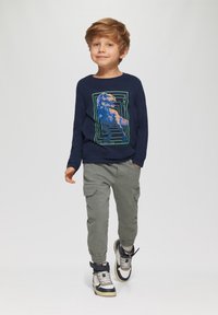 Marineblauw shirt met lange mouwen met een dinosauruslogo en groene accenten; olijfkleurige cargobroek met zakken; sneakers met zwarte en beige details.
