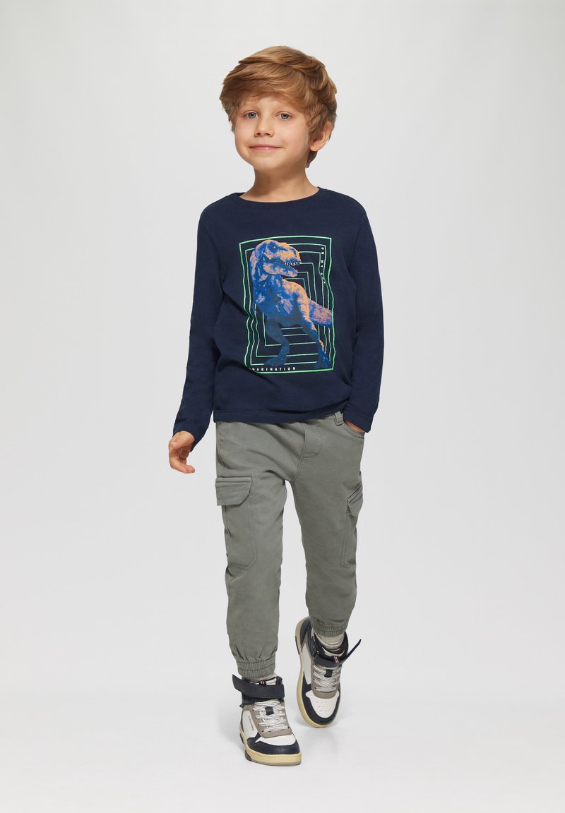 Marineblauw shirt met lange mouwen met een dinosauruslogo en groene accenten; olijfkleurige cargobroek met zakken; sneakers met zwarte en beige details.