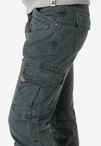 Cargo-Hosen aus dunkelgrüner Baumwolle mit mehreren Taschen, einschließlich einer Seitentasche, kontrastierenden Nähten und einem strukturierten Muster.