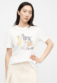 Γυναίκα που φοράει λευκό T-shirt με πολύχρωμους εικονογραφημένους σκύλους και το κείμενο "Barbour", συνδυασμένο με μια ανοιχτό μπεζ φούστα, ποζάρει με το χέρι στη μέση.