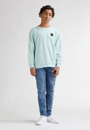 Lichtblauwe sweatshirt met een ronde hals en een klein zwart logoplaatje, gecombineerd met slim-fit blauwe jeans en witte sneakers.