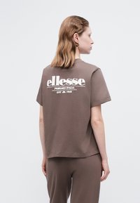 Sieviete ar plecu garumu matiem, tērpta brūnā ellesse t-kreklā un saskaņotās biksēs, stāvot sānis un rādot t-krekla aizmugurē esošo logotipu.