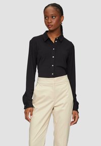 Black long-sleeve button-up shirt with a collar and cuffs, paired with light beige high-waisted trousers. Smooth fabric, minimalistic design.