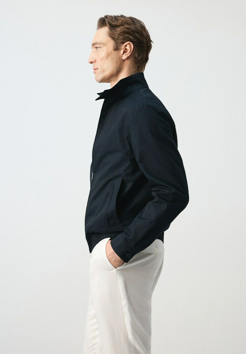 Homme en veste noire à col relevé et pantalon blanc, debout de profil, les mains dans les poches, sur fond uni.