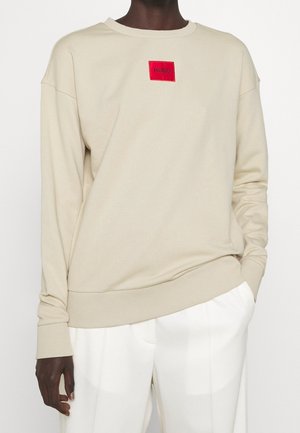 Beige sweatshirt met een ronde hals en verlaagde schouders, met een rode rechthoekige logo patch in het midden. Soepele textuur.