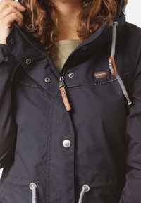 Tmavě modrá nepromokavá bunda se stříbrnými knoflíky, nastavitelnou kapucí a kontrastními šňůrkami; má přední zip a koženou značkovou visačku.