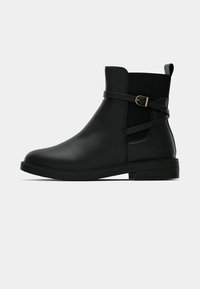 Črne gležnjenje z nizko peto, elastikami na straneh in dekorativnim pasom z zlatim sponko na enobarvni podlagi.