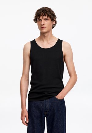 Jeune homme aux cheveux bruns bouclés portant un débardeur noir sans manches et un jean bleu foncé, debout avec une main dans la poche.
