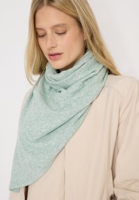 Donna bionda che indossa una morbida sciarpa triangolare di un leggero verde chiaro, drappeggiata attorno al collo sopra una giacca beige con zip.