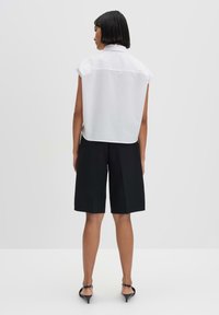 Blusa bianca senza maniche e pantaloni corti neri sartoriali. La blusa presenta un retro con yoke e un orlo curvo. I pantaloni corti sono lunghi fino al ginocchio con una vestibilità strutturata.