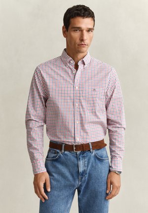 Mann trägt ein rot-blau-weiß kariertes Hemd mit Knöpfen, das in eine blaue Jeans mit braunem Gürtel gesteckt ist, und steht vor einem einfachen Hintergrund.