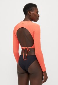 Coral long-sleeve swimsuit with textured fabric, open back design, and adjustable tie. Paired with a high-waisted black bikini bottom.