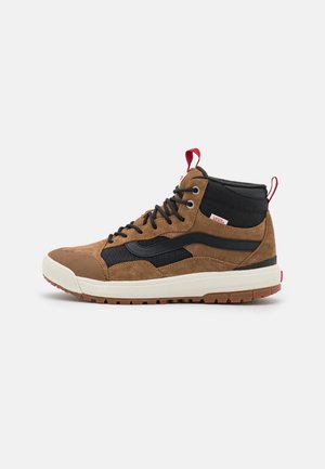 Bruine suède en zwarte stoffen hoge sneakers met een witte tussenzool, rubberen buitenzool en een rood treklipje bij de hiel.