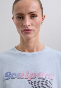 Junge Frau mit hochgestecktem Haar, die ein hellblaues Scalpers-T-Shirt mit pink- und lila-farbenem Farbverlauf und einem karierten Flaggenmuster trägt.