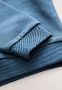 Poignet de sweat-shirt bleu avec un tissu lisse à texture côtelée. Le design présente un bord plié et une couleur uniforme sur toute la surface.