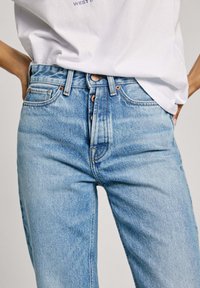 Vaqueros de denim azul claro con una cintura alta, cinco bolsillos y herrajes de cobre. La tela tiene una textura ligeramente desgastada.