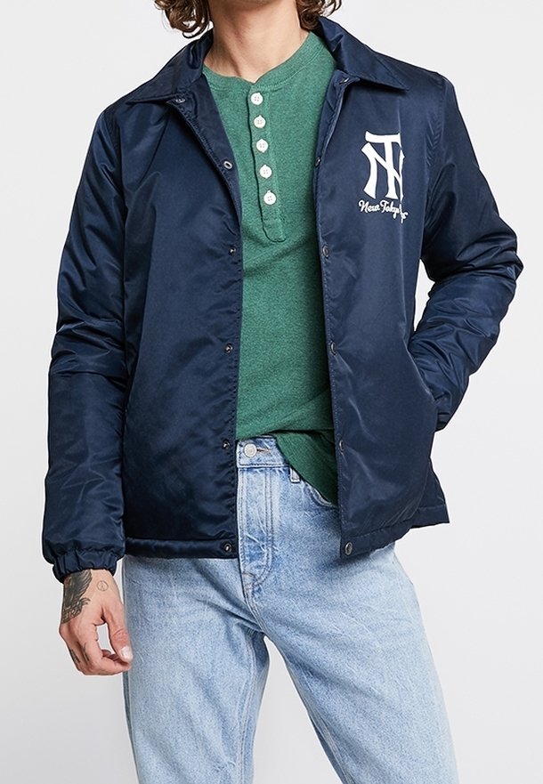 Mann trägt eine marineblaue, geknöpfte Jacke mit weißem Logo über einem grünen Henley-Shirt und hellblauen Jeans, Hand in der Tasche, tätowiert sichtbar.