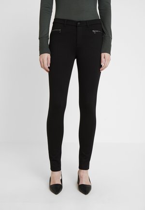 Femme portant un pantalon noir ajusté avec des poches zippées à l'avant, assorti à un haut sombre à manches longues et des talons noirs à bout pointu.