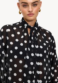 Blouse noire transparente dotée d'un col noué et de grands pois blancs. Manches longues avec poignets boutonnés. Tissu léger et fluide.