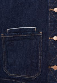 Gros plan d'un tissu en denim bleu foncé montrant une poche avant avec des surpiqûres dorées et des boutons métalliques sur le côté droit.