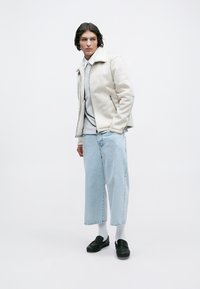 Giovane che indossa un giubbotto in shearling crema, camicia grigio chiaro, jeans azzurri cropped, calze bianche e mocassini neri, in piedi su uno sfondo bianco.