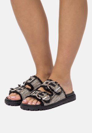 Pies descalzos que llevan sandalias tipo slide con textura en blanco y negro, suelas gruesas y hebillas grandes de plata en dos correas.