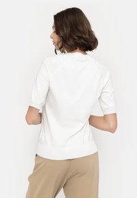 White short-sleeved top with a round neckline, ribbed cuffs and hem, and a smooth texture. Worn with beige pants, shown from the back.