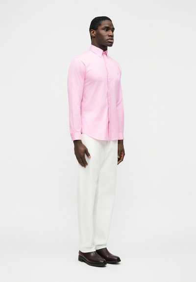 Muž stojaci v bielych nohaviciach, ružovej košeli na gombíky a tmavohnedých topánkach na jednoduchom svetlom pozadí.