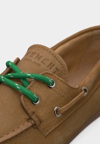 Καφέ παπούτσι σκάφους από σουέντ με πράσινα κορδόνια περασμένα μέσα από ασημένιες οπές, με ανάγλυφο λογότυπο στη γλώσσα.