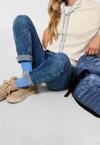 Beige hoodie med ljusblå snören, ljusblå strumpor, blå jeans och beige sneakers. En silverfärgad puffjacka ligger i närheten. Mjuka texturer synliga.