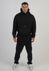 Conjunto de sudadera y pantalones de chándal negros, hecho de algodón. La sudadera cuenta con un bolsillo frontal y un logotipo, mientras que los pantalones tienen un logotipo sutil cerca del dobladillo.
