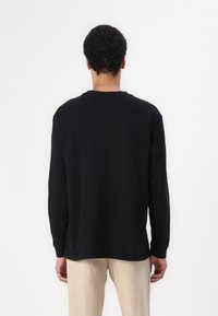 Black long-sleeve shirt with a crew neck, soft texture, dropped shoulders, and a relaxed fit, worn with light-colored, loose-fitting trousers.