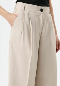 Beige, hooggetailleerde broek gemaakt van gestructureerde stof. Voorzien van een enkele knoopsluiting, plooien en steekzakken aan de zijkant.