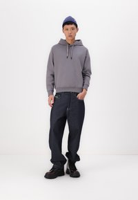 Sudadera gris con capucha y cordones, combinada con unos jeans de mezclilla oscuros de corte holgado y botas negras de suela gruesa, sobre un fondo blanco.